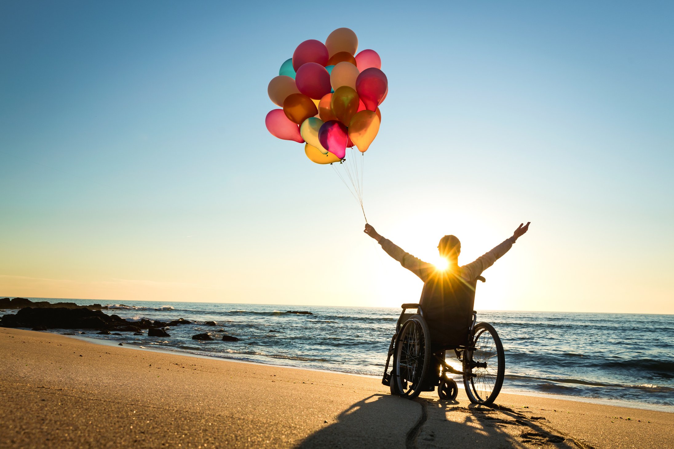 a person in a wheelchair facing towards the sun holding balloons on the beach