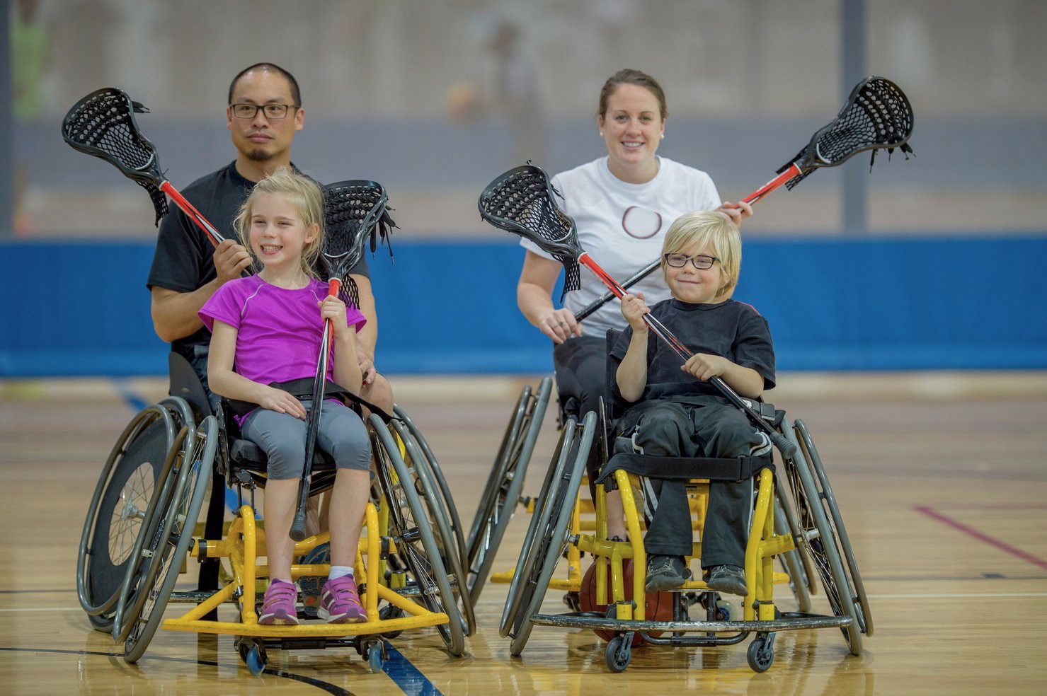 four people in wheelchairs holding lacrosse sticks. The two people in the front are children and the two people in the back are adults. They are smiling and appear happy. 