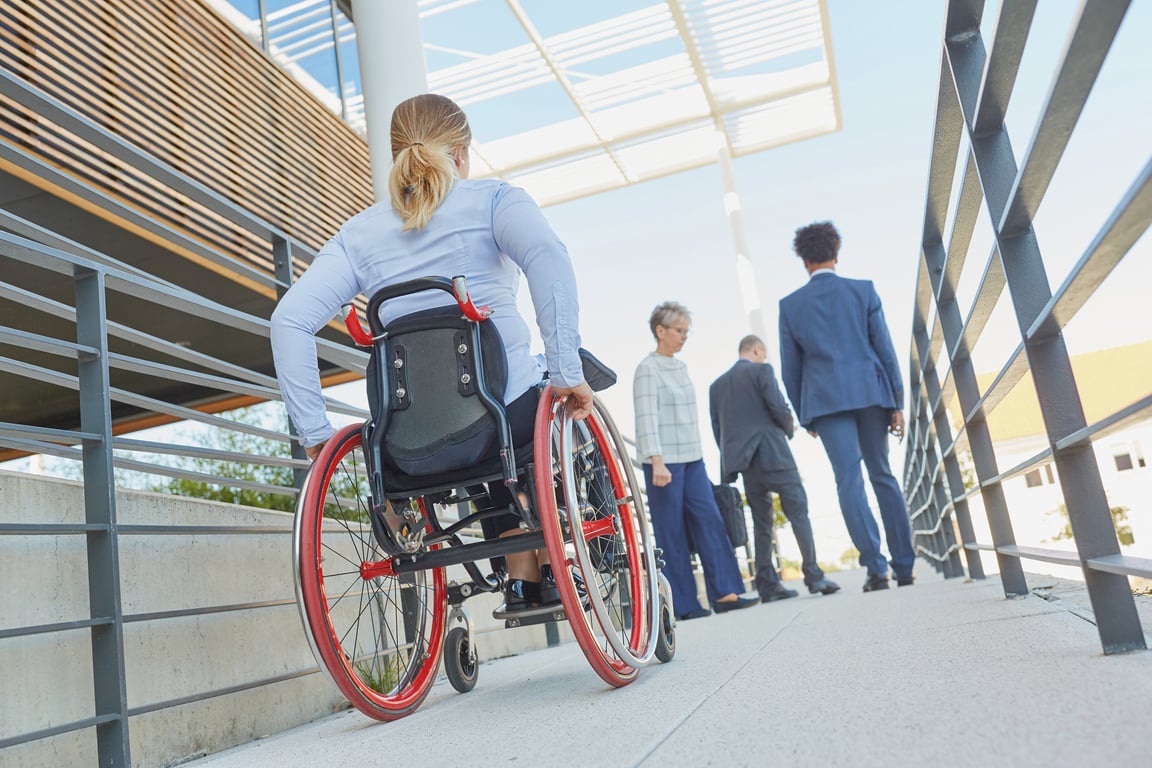 Person in a blue shirt sitting in a wheelchair pushing up a ramp towards a building. Three additional people in business clothes are walking up the ramp towards a building. 