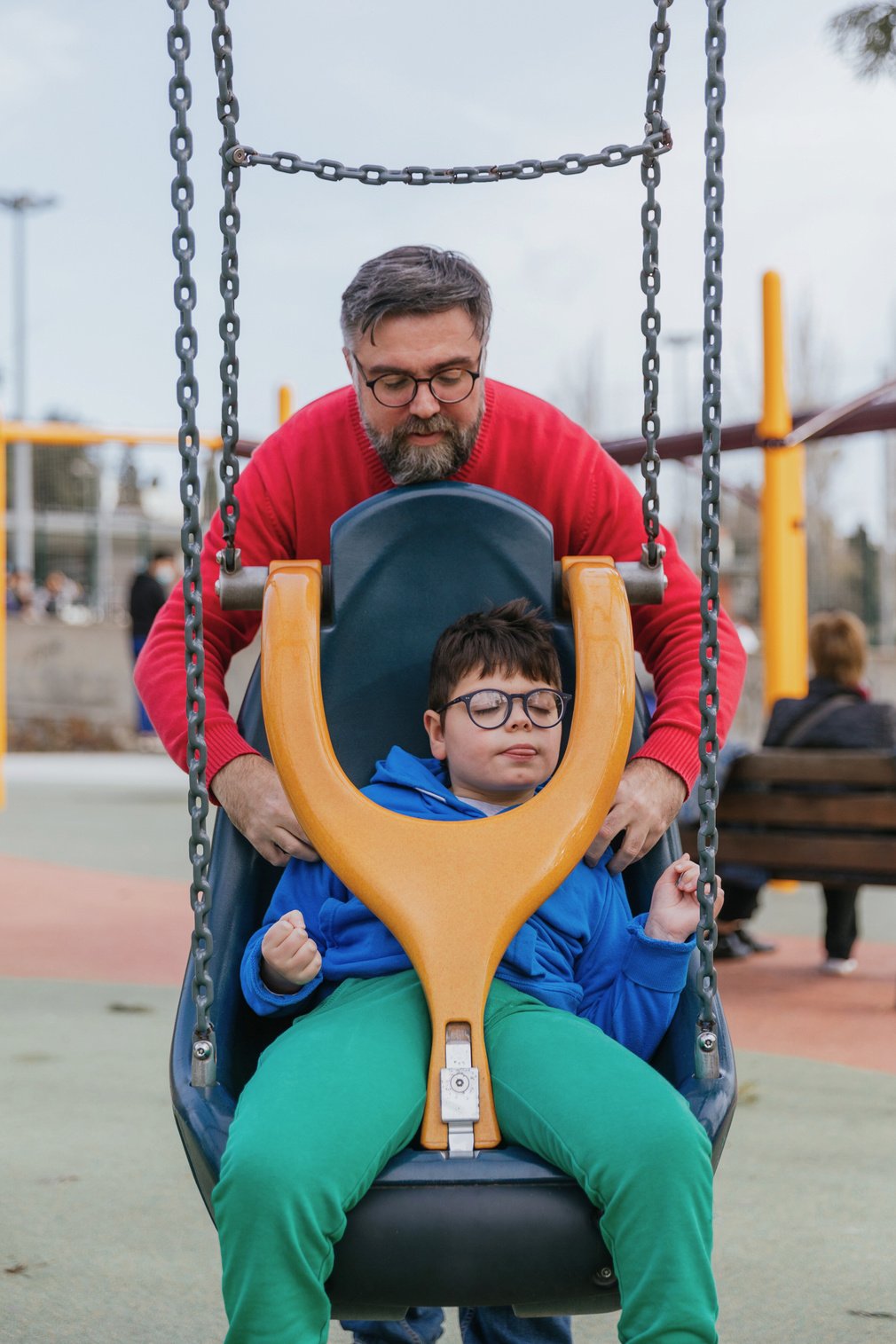 a person in a red sweater wearing glasses and a beard standing behind an accessible swing. A child with glasses and a blue jacket is sitting in the swing. The swing has a yellow Y-shaped harness that helps with stabilization.